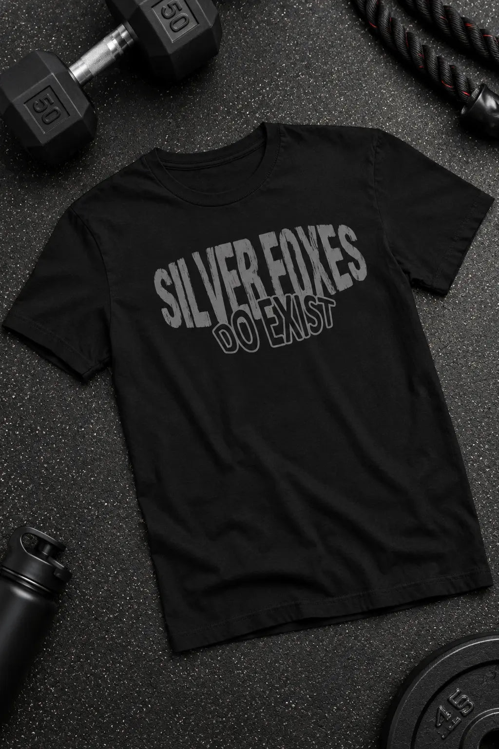 A black short-sleeve graphic t-shirt laid flat on a dark gym floor, surrounded by dumbbells, a rope, and a water bottle. The shirt features the text 'SILVER FOXES DO EMST' in distressed gray lettering.