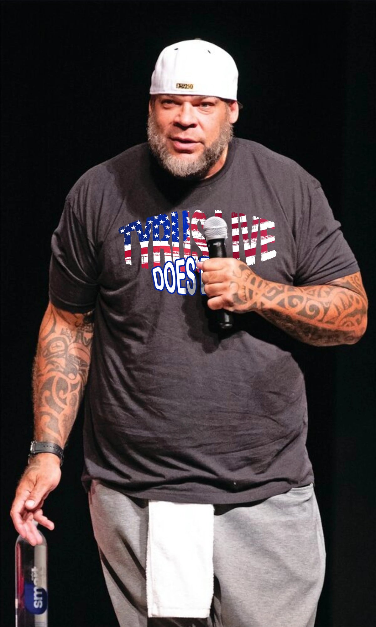 Tyrus wearing a black "Tyrus Live Does Exist" shirt, white cap, and gray sweatpants, holding a microphone on a stage.