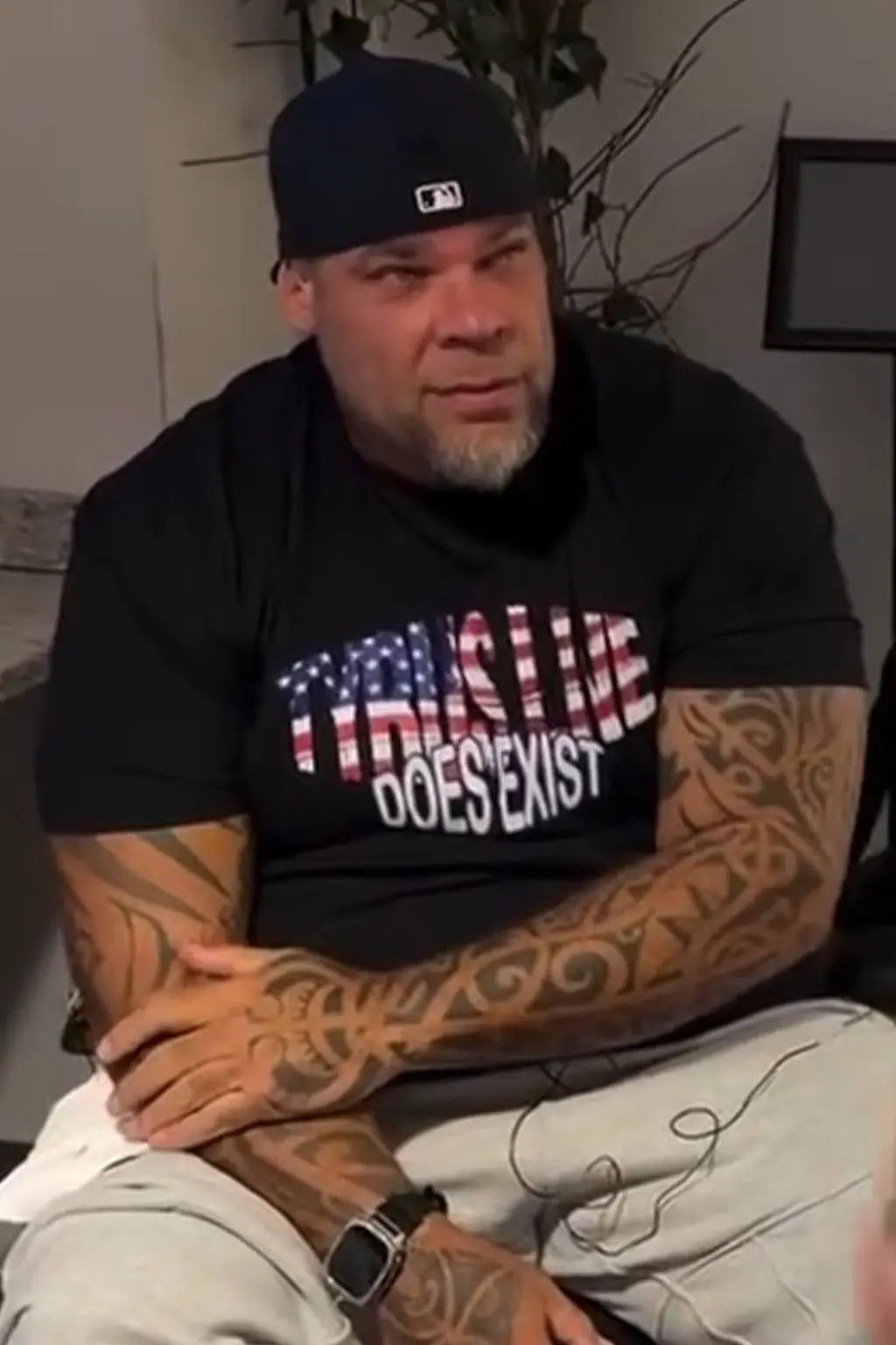 A muscular man with tattoos wearing a black t-shirt with the text 'TYRUS LIVE DOES EXIST' in an American flag pattern, a black baseball cap, and light gray sweatpants, sitting indoors.