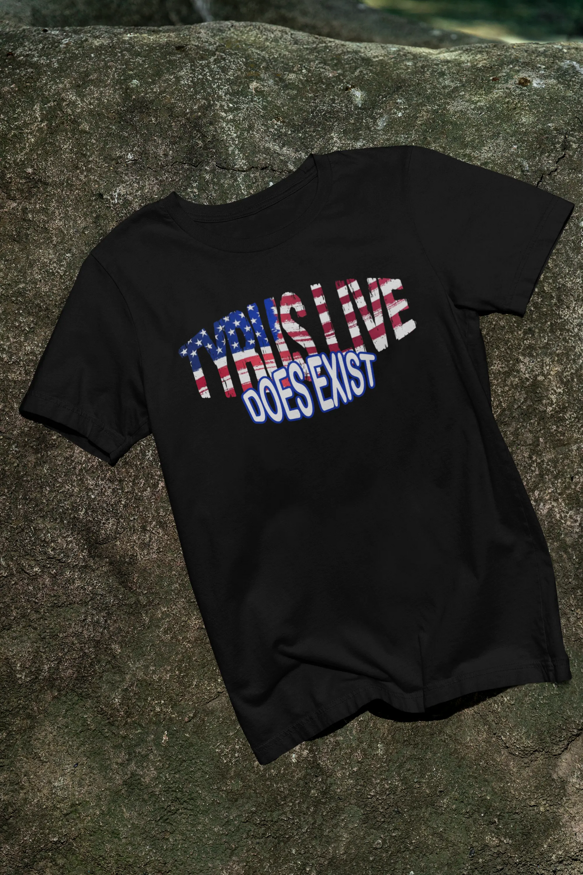 A black short-sleeve t-shirt laid flat on a textured rock surface. The shirt features a graphic print on the front reading 'TYRUS LIVE DOES EXIST' in stylized lettering, with the first word filled with an American flag pattern and the second word in blue with a white outline.